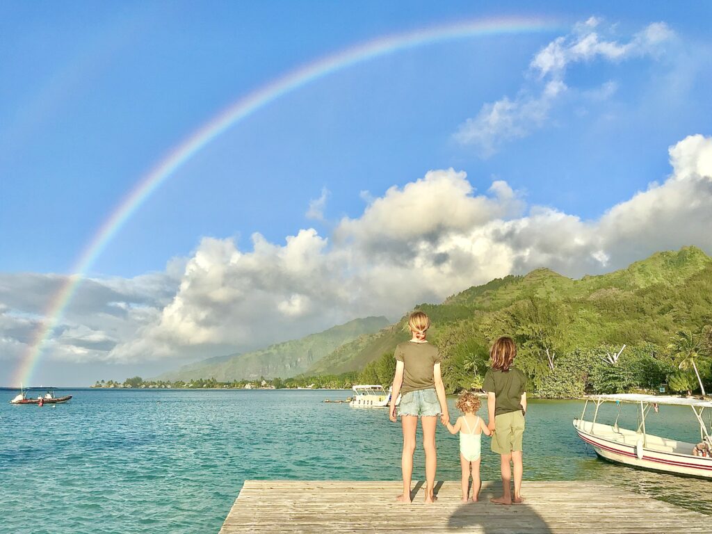 Three kids stand on the edge of a dock looking at a rainbow over blue water and clouds.